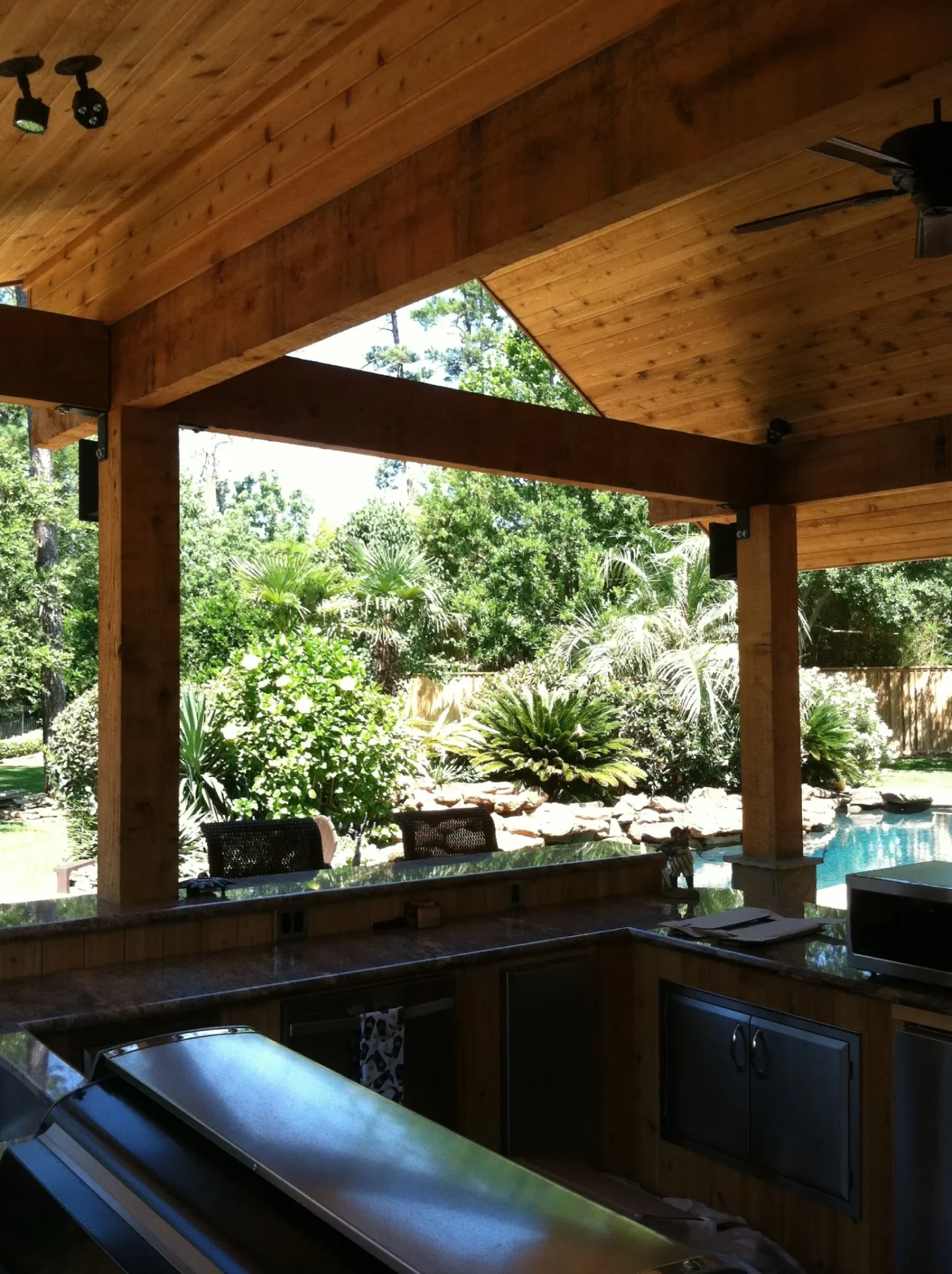 An outdoor kitchen under a wooden roof overlooks a backyard with lush green plants and a pool. The kitchen features a sink, counter space, and stainless steel appliances.