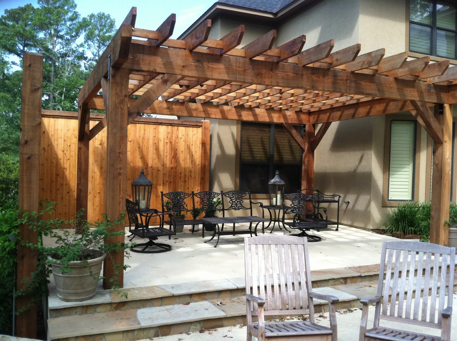 A backyard patio features a wooden pergola with a lattice roof, wrought iron furniture, a potted plant, and a wooden privacy fence, adjacent to a house with beige walls and double-hung windows.