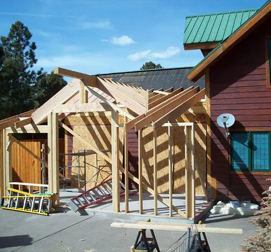 A house under construction with a partially built wooden framework extending from the existing structure. A green ladder and construction materials are visible on the ground. Trees and sky are in the background.