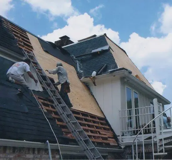 Two workers on ladders repair a steeply sloped roof of a house under a blue sky with clouds.