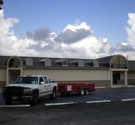 A white truck with a red trailer is parked in front of a building with multiple arched windows under a partly cloudy sky.