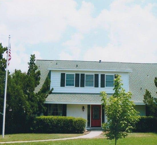 A two-story white house with green shutters and a red front door, surrounded by green bushes and trees, and an American flag on a pole in the front yard.